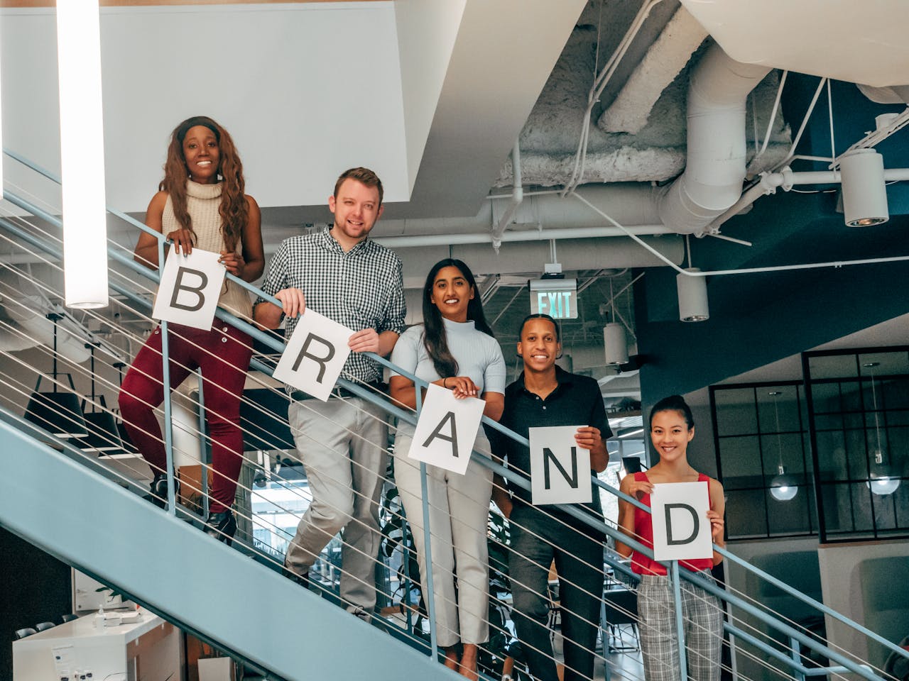 Diverse group of adults smiling and holding 'BRAND' signs on an office staircase.
